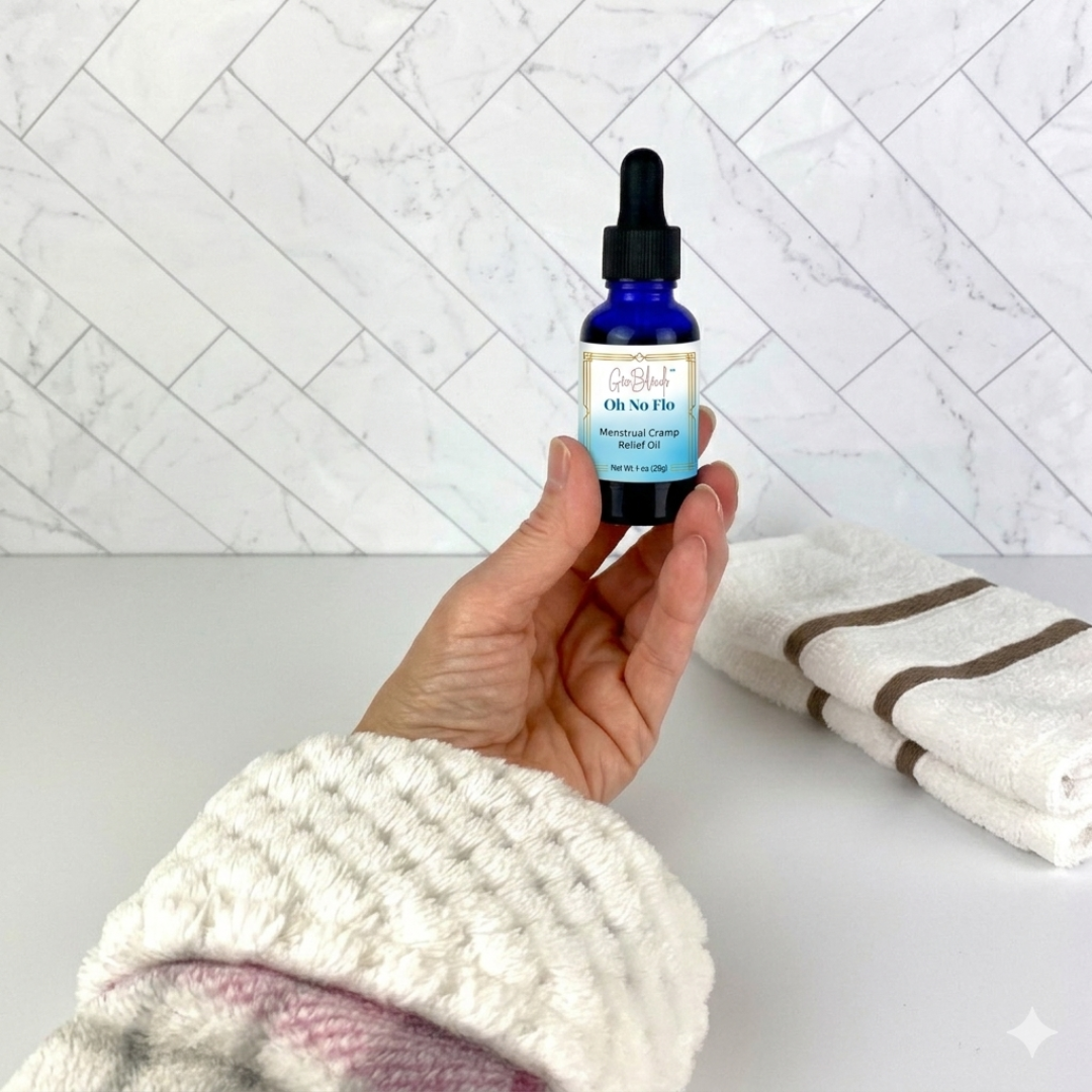 Hand holding a bottle of 'Oh No Flo Menstrual Cramp Soothing Oil' with a dropper against a white marble background